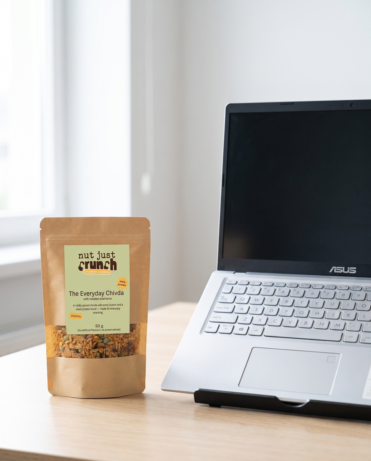 A 50 g Edamame Chivda pouch on a work desk beside a laptop